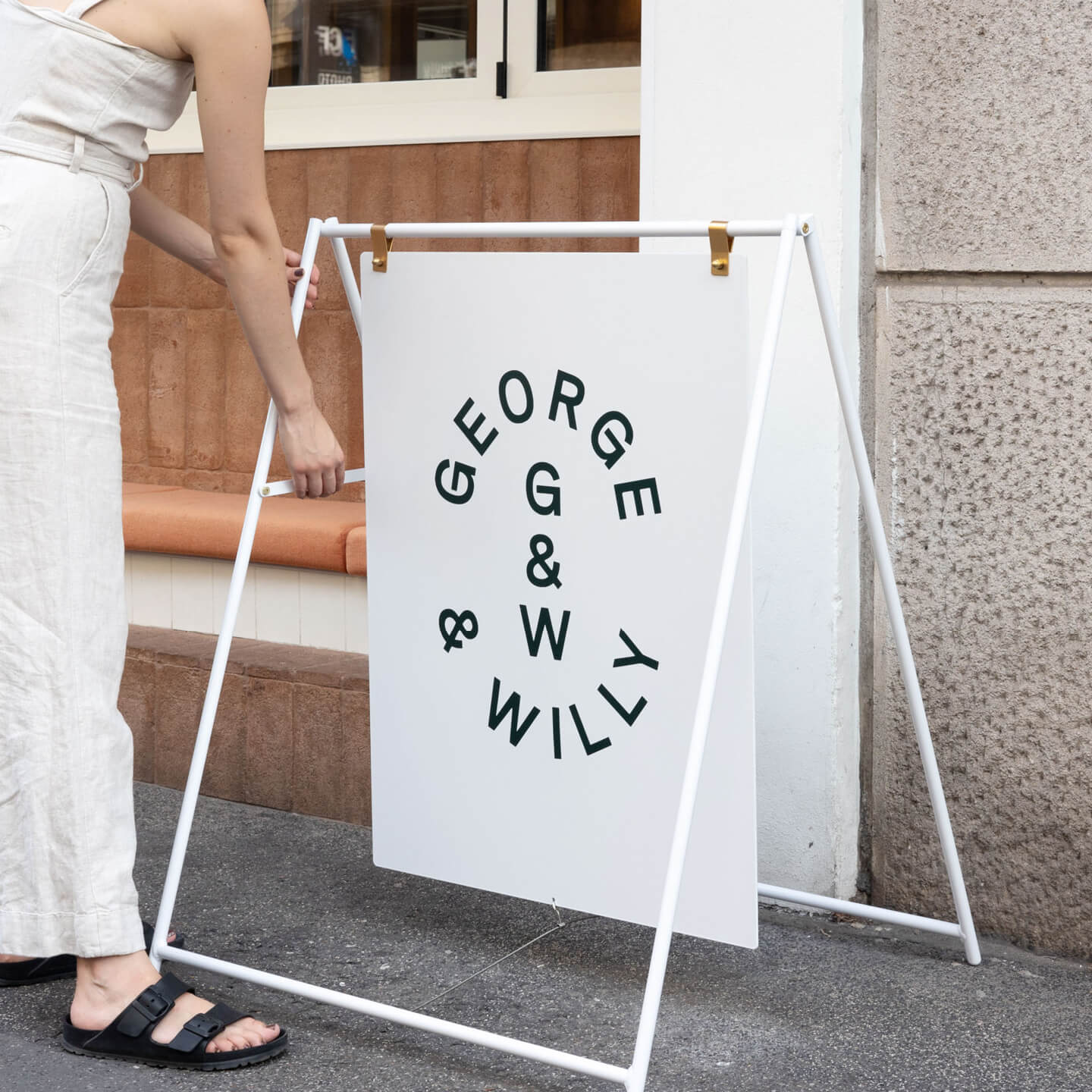 white large sidewalk sign on sidewalk with george & willy logo with person adjusting the frame