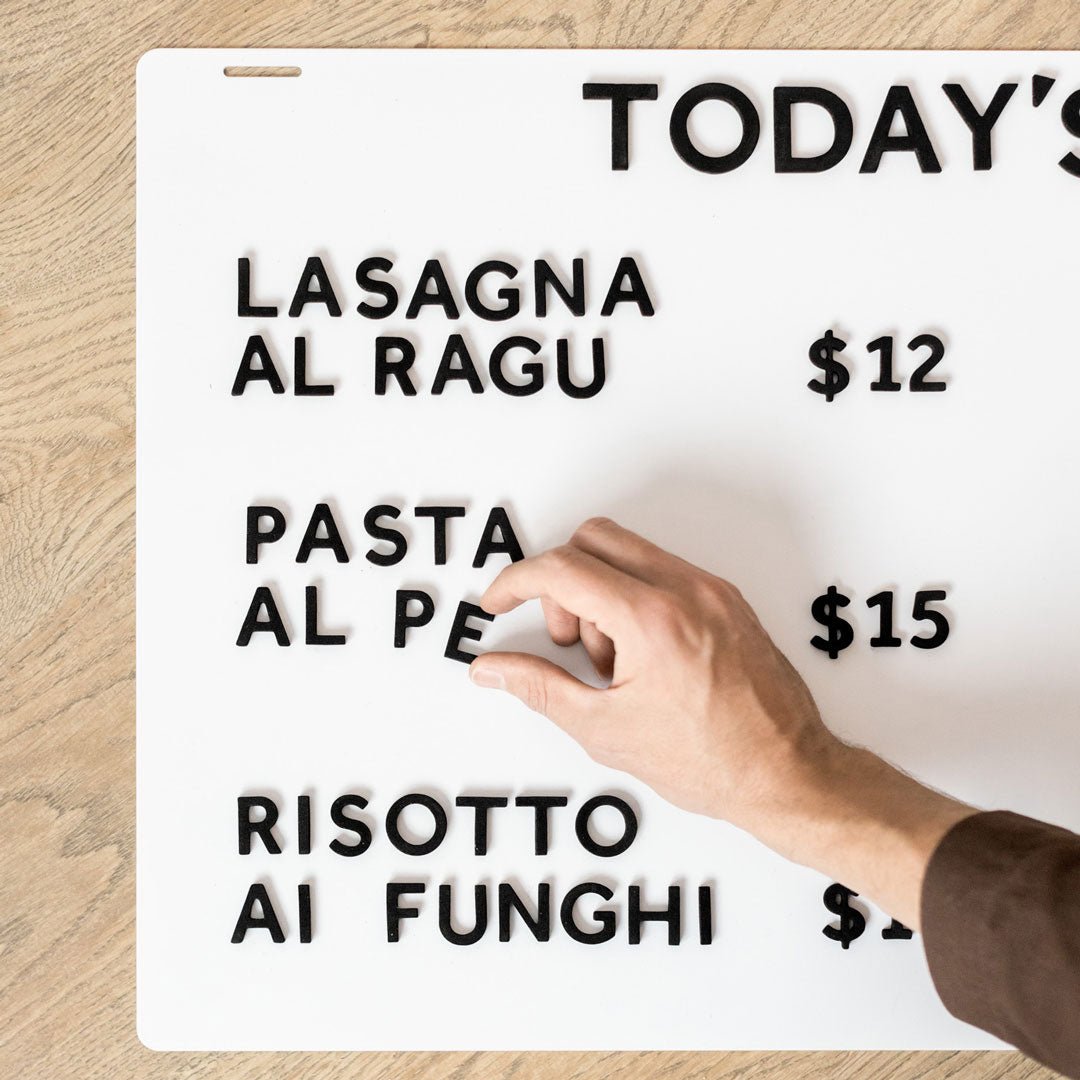 hand applying black magnet letter to a white menu board displaying pasta