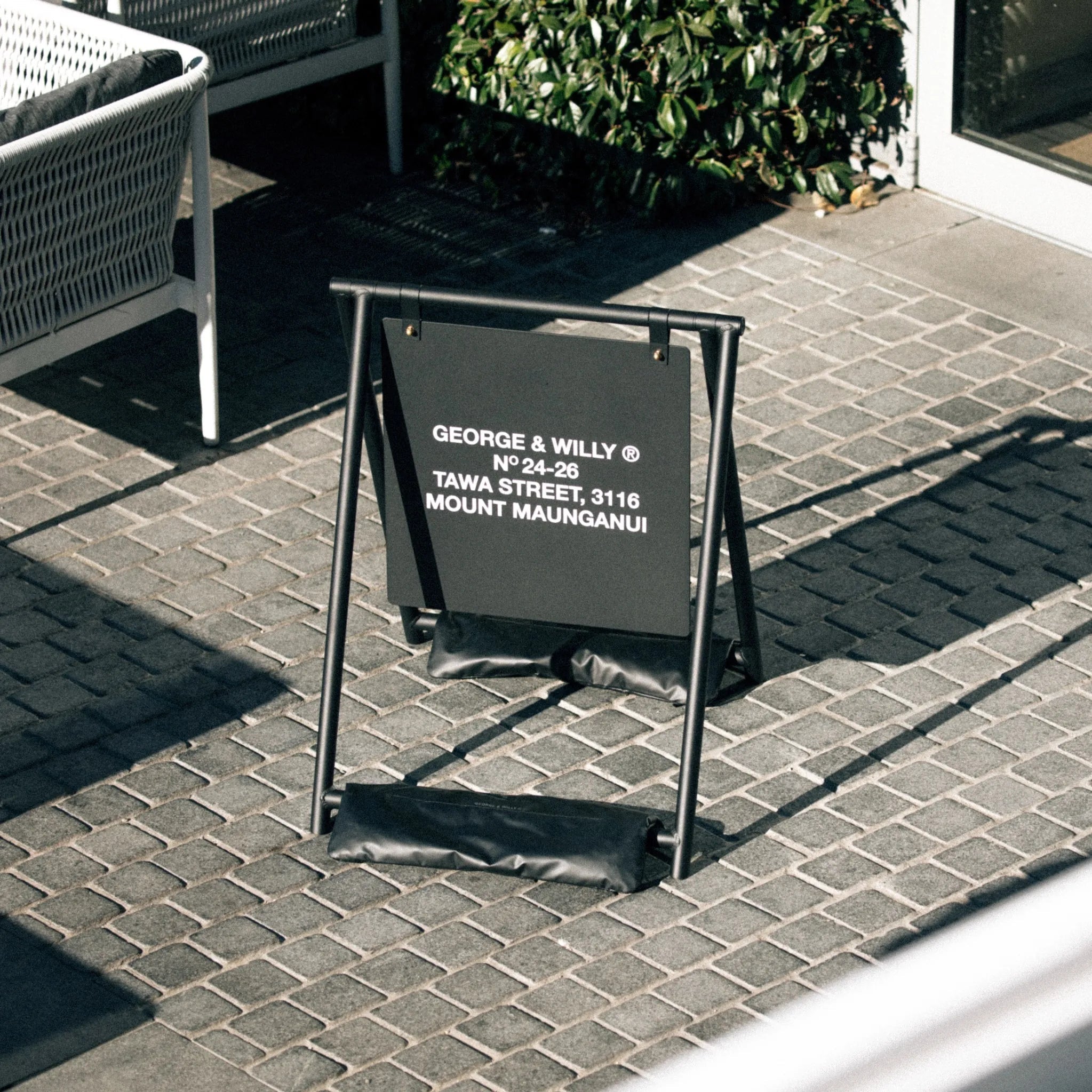 large sandwich board style sidewalk business sign with red logo on it using black sandbags