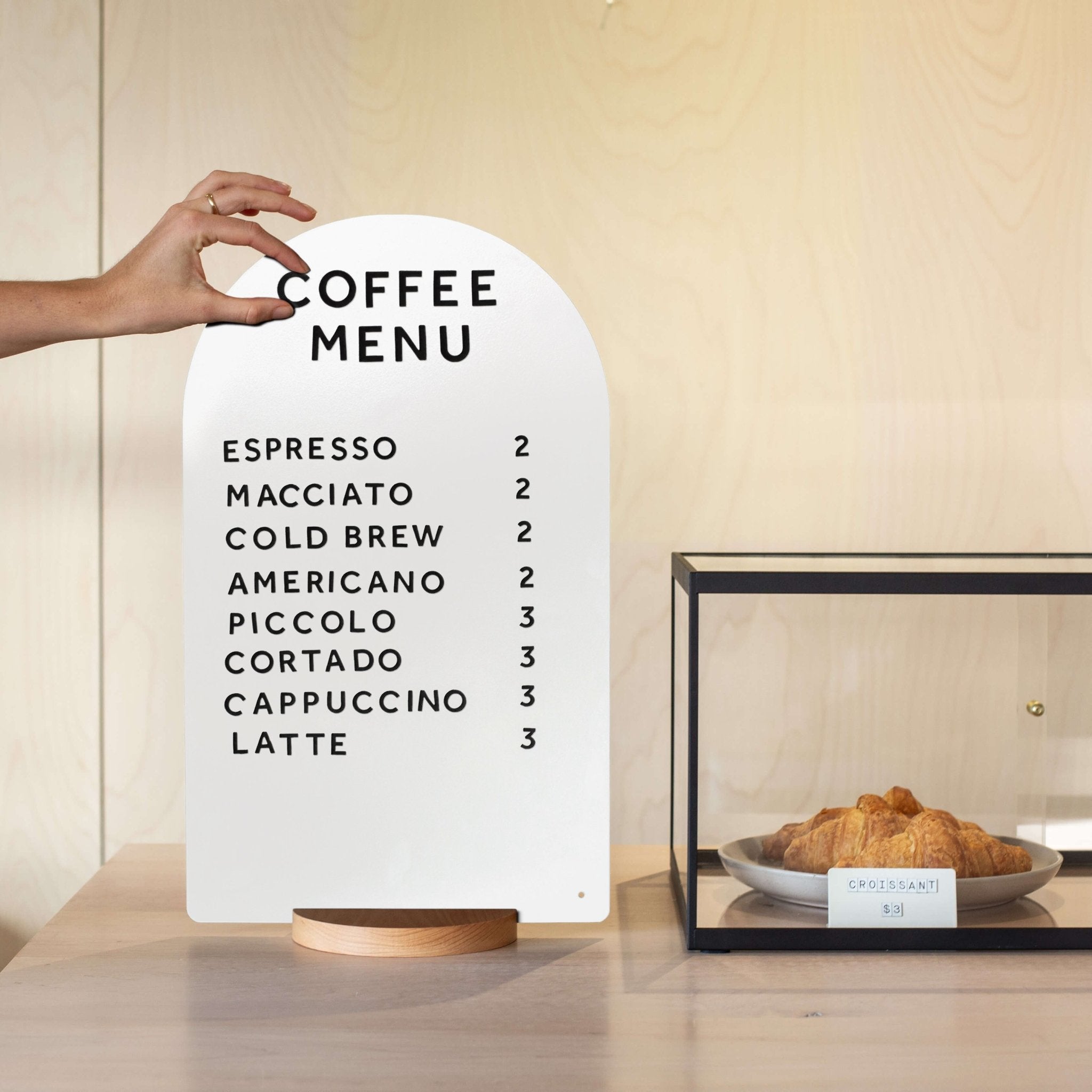 close up of hand changing magnet letters on a white arch tabletop sign, beside a bakery display case